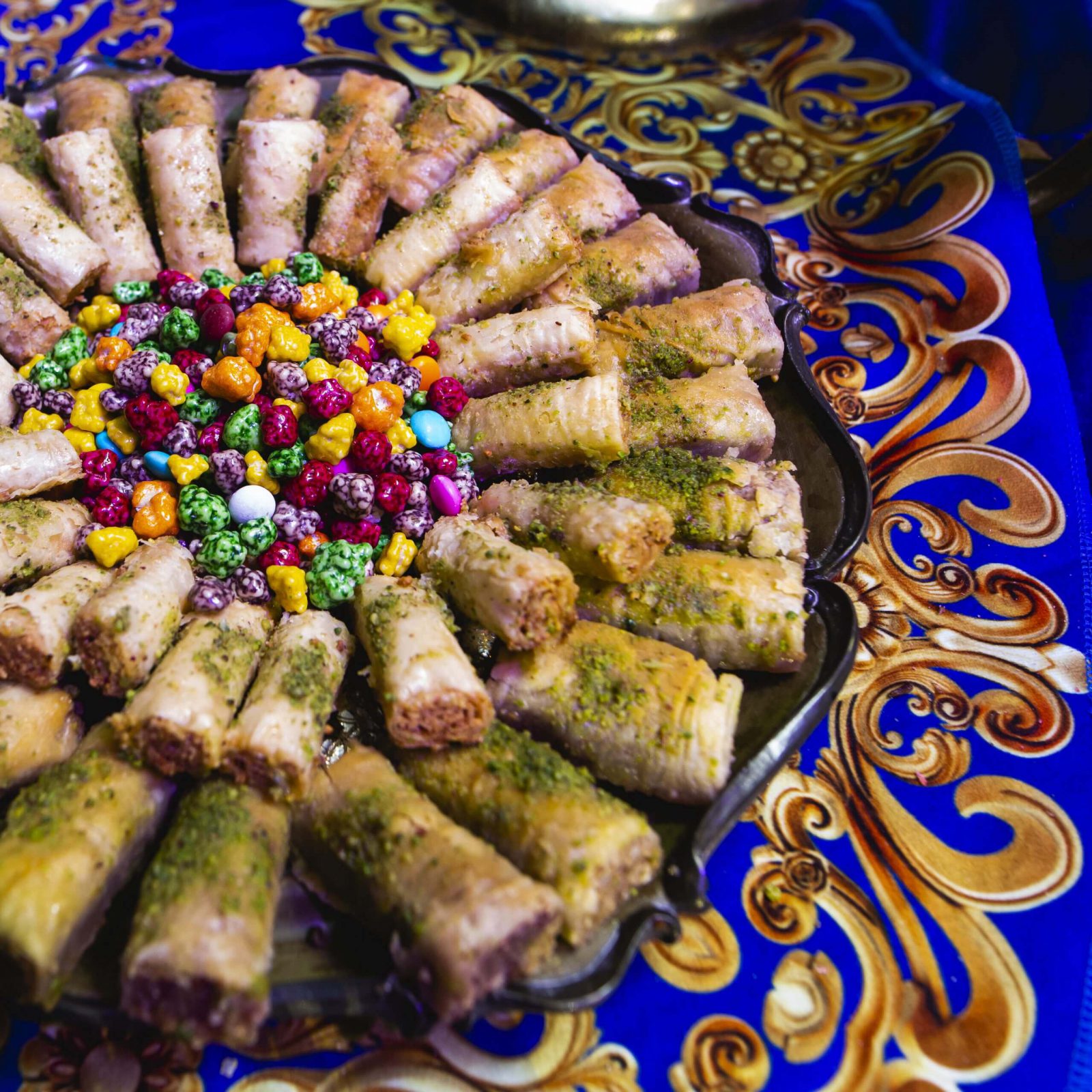 מגש מתוקים צבעוניים עם קינוחים, ממתקים ופסי בקלוואה, מוגש על רקע טקסטיל מעוצב.