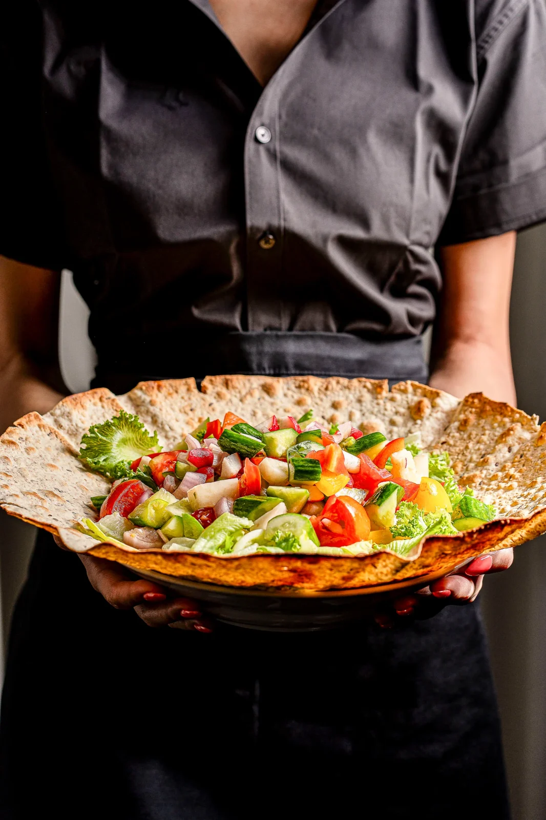 צלחת עם סלט צבעוני על פיתה גדולה, מוגשת על ידי אדם בגד כהה.