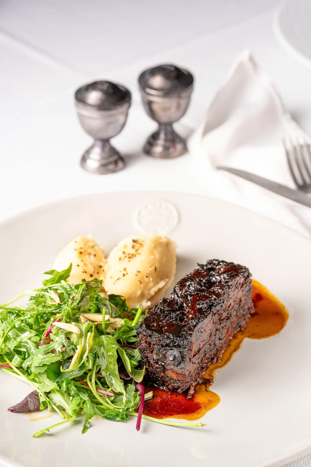 מנה מעוצבת עם בשר, סלט ירוק, ופירה, לצד גביעים ממתכת על שולחן מוכן לאירועים.