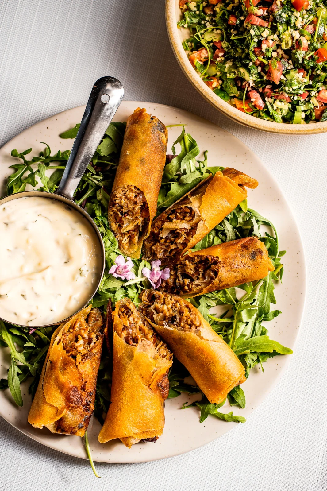 צלחת עם רולים מטוגנים על עלי רוקט, ליד רוטב סמיך וסלט ירקות צבעוני.