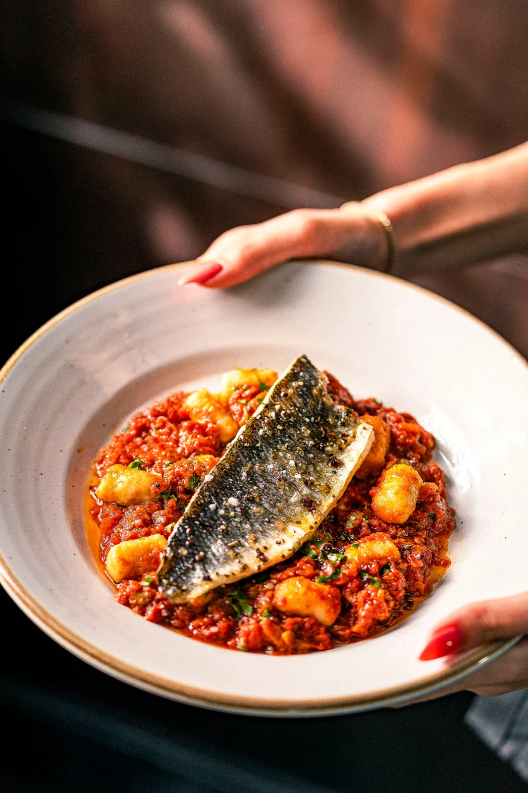 צלחת עם פילה דג על רוטב עגבניות, עם פסטה תפוחי אדמה, מוגשת ביד.