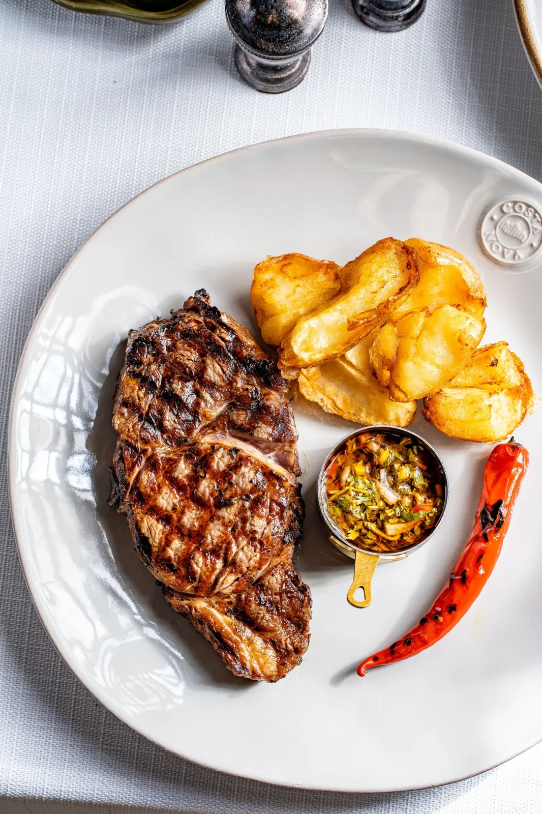 צלחת עם סטייק צלוי, תפוחי אדמה צלויים ורוטב עם ירקות, מוגשת על רקע שולחן מסודר.