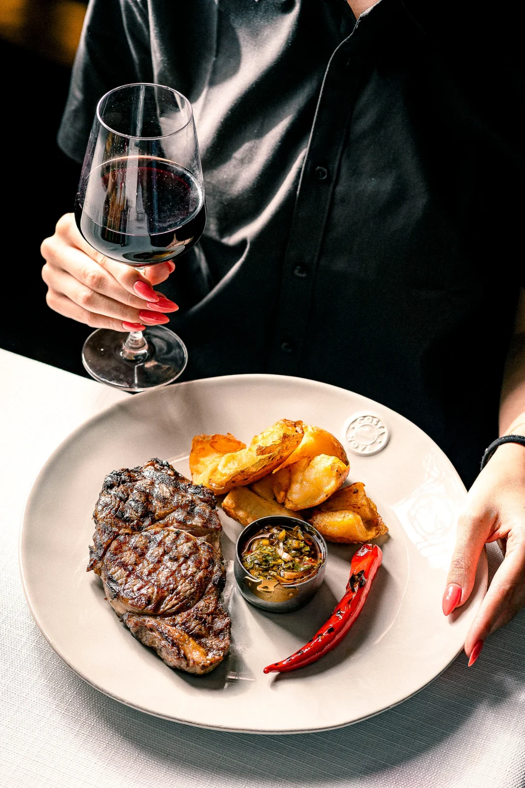 מנה של סטייק עם תוספת תפוחי אדמה, רוטב חריף וכוס יין אדום.
