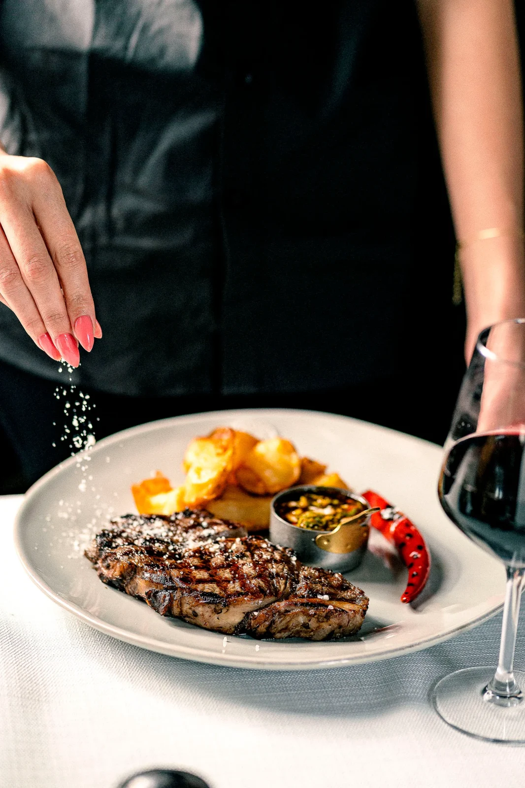 צלחת עם סטייק צלוי, תפוחי אדמה ויין אדום, יד מפזרת מלח מעל המנה.