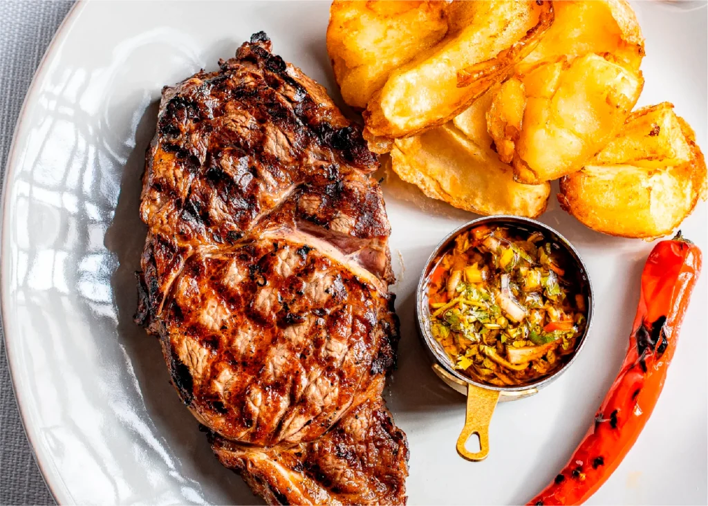 צלחת עם סטייק צלוי, תפוחי אדמה מטוגנים, רוטב ירוק ופלפל חריף אדום.