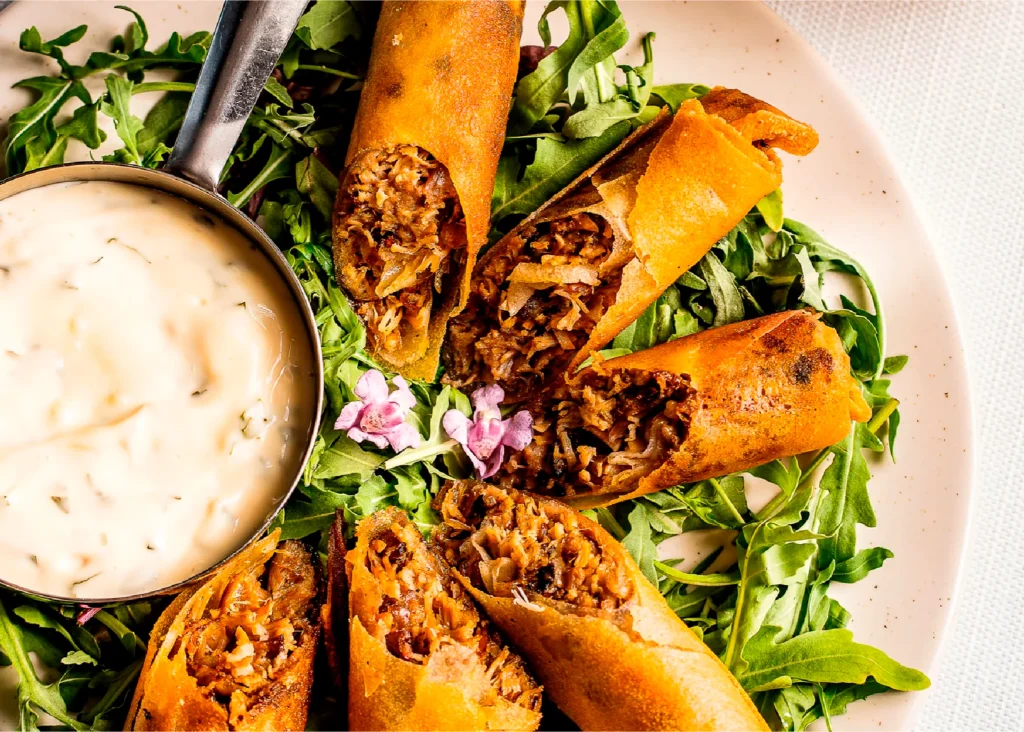 מגש עם רולס מטוגנים ממולאים בירקות, מונחים על עלי רוקט עם רוטב ליד.